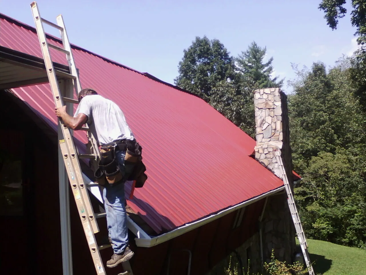 Roofer on a ladder beside a red metal roof and stone chimney — Soffit & Fascia Repair by Roofing Brothers in Lake Dallas
